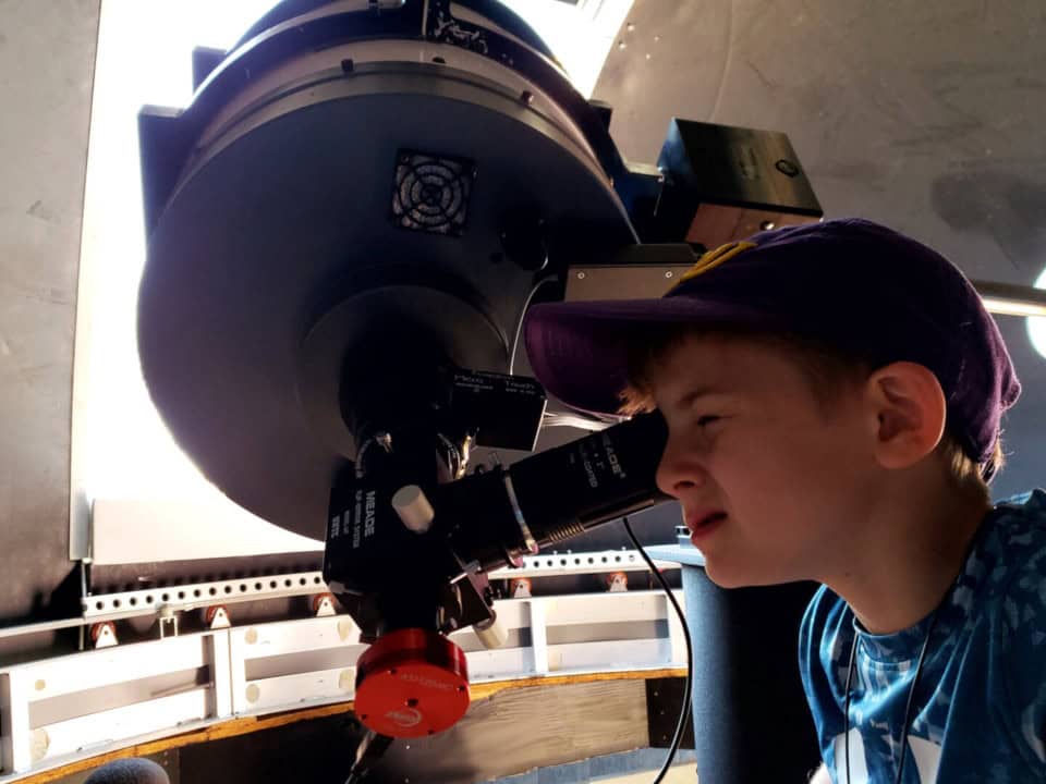 Boy in a purple ECU hat looks through a telescope at the daytime moon.